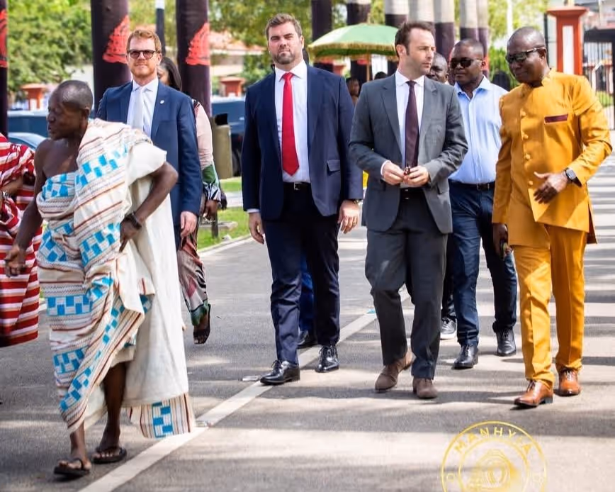 Group of men walking outdoors on a sunny day, including two men in traditional African cloth and four men in formal suits and business attire.